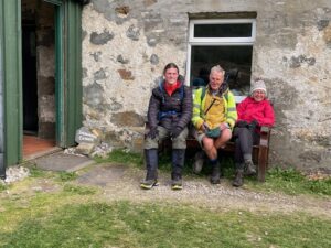 Three people sit on a bench next to a stone wall